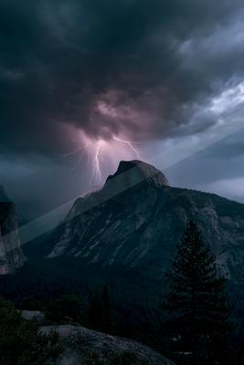 Intense lightning bolt strikes the summit of a rocky mountain peak beneath a dark, turbulent storm cloud, with a dense evergreen forest in the foreground.