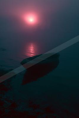 A small wooden boat rests on the shore of a calm lake enveloped in thick fog, with a glowing red sun rising in the background and casting a vibrant reflection o