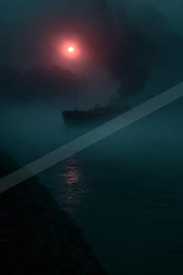 A large ship moves through thick fog on a river, with a glowing red sun in the hazy sky and its reflection shimmering on the dark water.