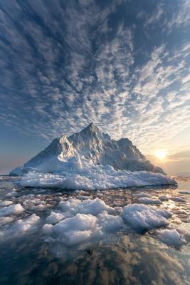 A large white iceberg floats in a polar sea filled with smaller ice chunks under a dramatic cloudy sky illuminated by the setting sun.