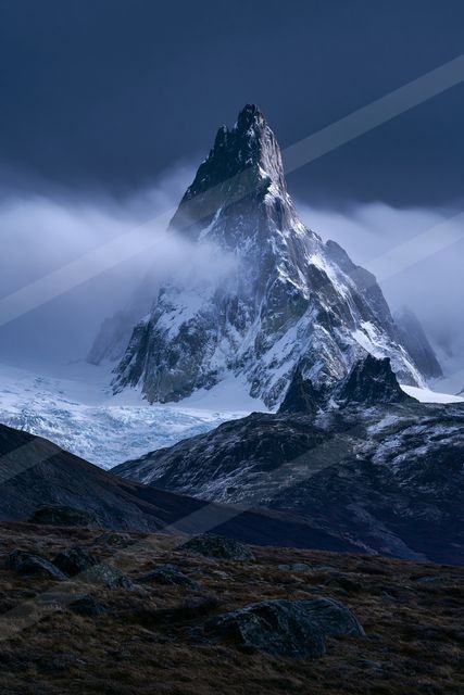 A towering snow-capped mountain peak piercing through a layer of clouds, with a glacier in the background and rocky foreground under twilight lighting.