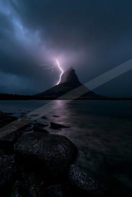 A powerful lightning bolt strikes the summit of a dark mountain peak during a thunderstorm, its light reflecting on the calm surface of a lake in the foreground