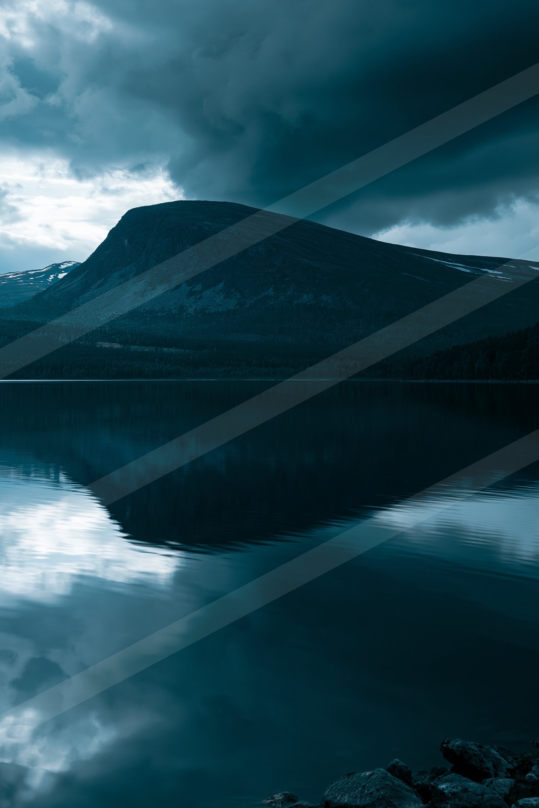 A serene twilight scene featuring a mountain peak with patches of snow, reflected perfectly in the calm waters below. The sky is filled with dark clouds, creati