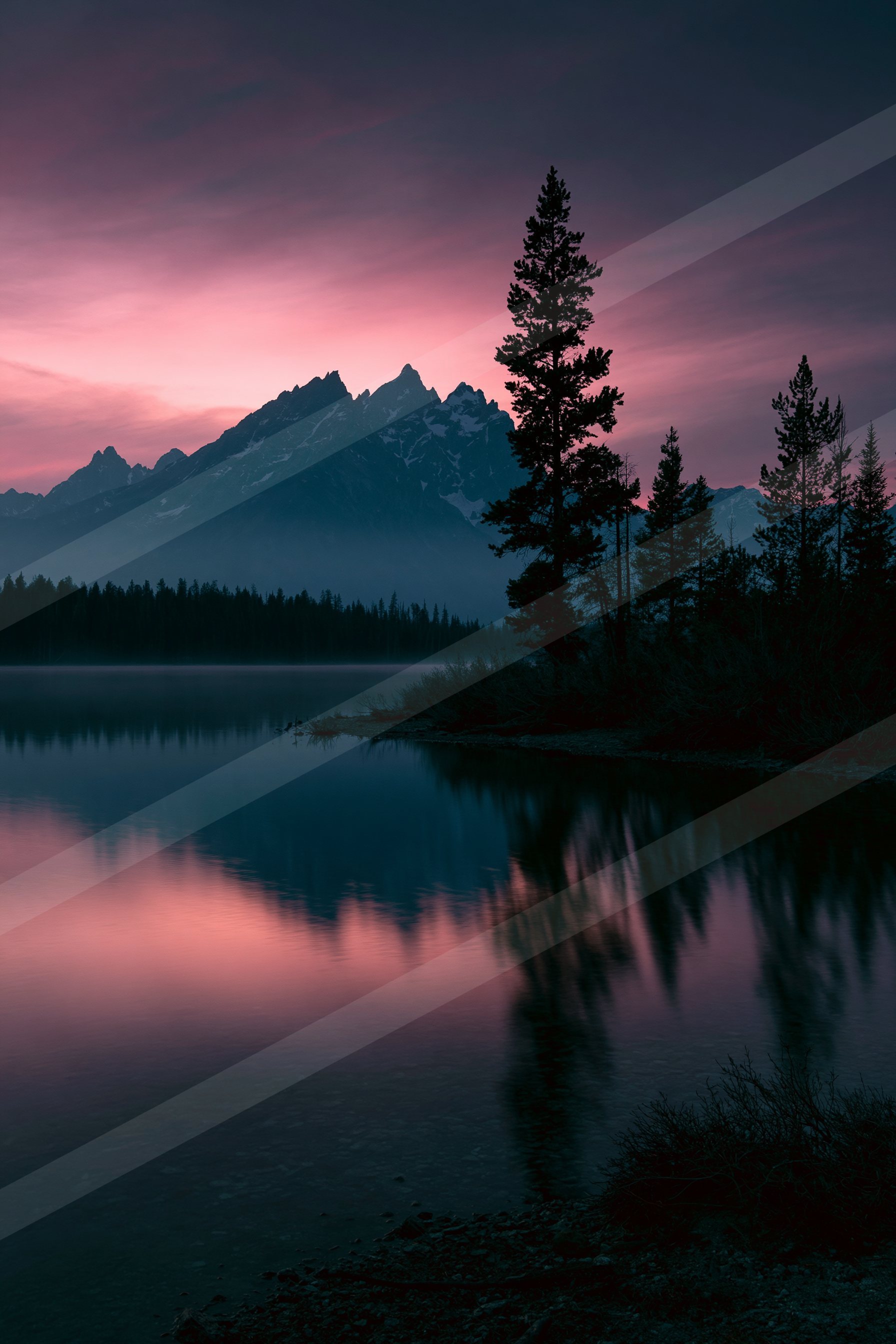 A serene lake reflects the silhouettes of trees and distant mountains under a vibrant twilight sky.