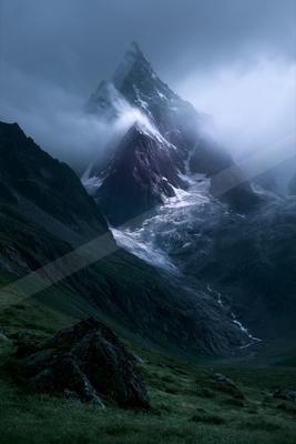 A sharp, snow-covered mountain peak rises through thick clouds and mist, with a glacier flowing down its slopes into a green valley in the foreground.