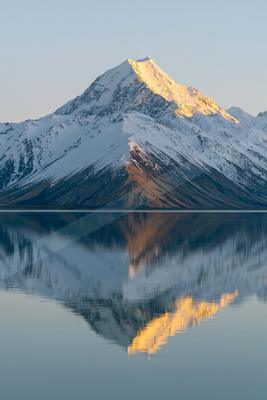 A majestic snow-covered mountain peak illuminated by golden sunrise light, perfectly mirrored in the still surface of a foreground lake.