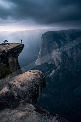 A lone individual stands at the edge of a rocky cliff, overlooking a vast canyon shrouded in mist and storm clouds.