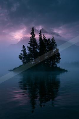 Silhouetted evergreen trees on a small island in a calm lake, with dramatic purple and pink twilight sky, mist, and mountain range in the background, their refl