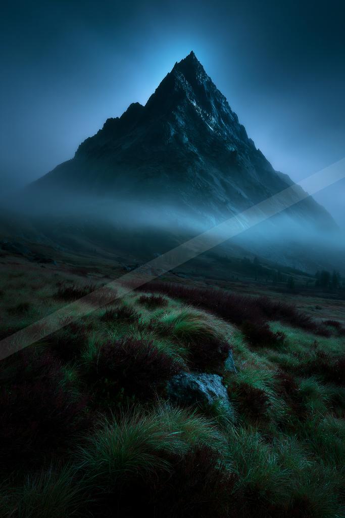 A sharp rocky mountain peak rises above thick blue mist in a dramatic twilight landscape, with a foreground of green and reddish heather and grass.