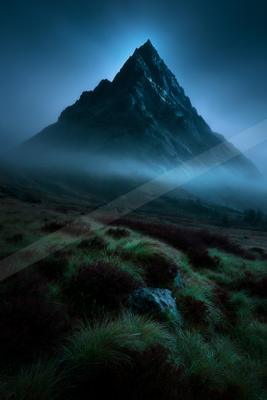 A sharp rocky mountain peak rises above thick blue mist in a dramatic twilight landscape, with a foreground of green and reddish heather and grass.