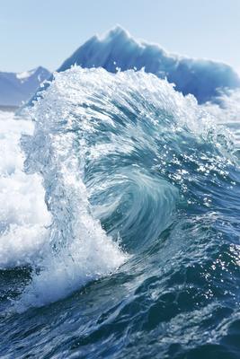 A powerful blue ocean wave crests and crashes with white foam and spray in the foreground, with a large blue iceberg visible in the background under a clear sky