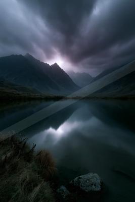 A serene mountain lake perfectly reflects the dark, stormy clouds and jagged peaks above, with a bright break in the clouds creating a glowing reflection on the