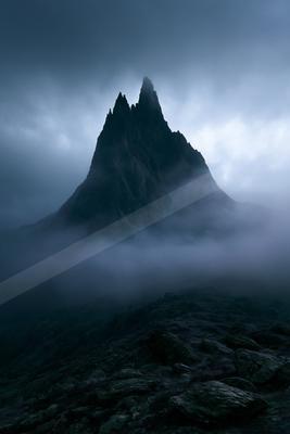 A sharp rocky mountain peak emerges from thick white fog and dark storm clouds, with a rocky foreground under a moody sky.