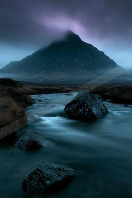 A fast-flowing river with large dark rocks in the foreground winds through a rugged landscape toward a massive mountain partially shrouded in thick mist under a