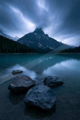 A tranquil lake mirrors a towering mountain peak under a moody sky.