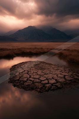 A stark desert landscape featuring a cracked earth mound surrounded by a reflective pool, set against a backdrop of stormy skies and distant mountains.