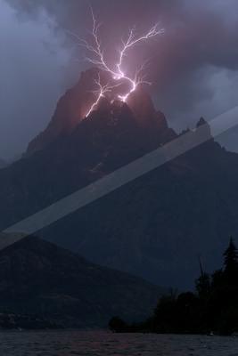 Dramatic lightning bolt strikes the summit of a rugged mountain peak during a thunderstorm, illuminating the rocky slopes above a dark lake with forested shorel