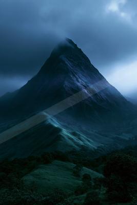 A sharp rocky mountain peak rises through thick fog and dark clouds under a deep blue twilight sky, with rolling green hills and trees in the foreground.