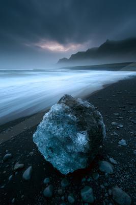 A large translucent blue ice boulder rests on a dark pebble beach as smooth white waves from the ocean flow past under a dramatic cloudy sky with distant misty 
