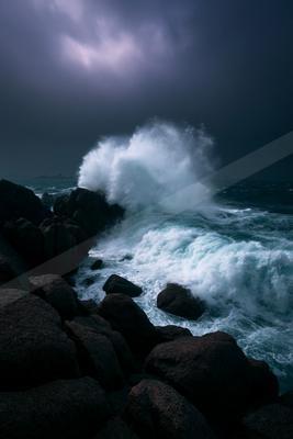 Powerful white wave explodes against jagged dark rocks under a moody, overcast sky in a stormy seascape.