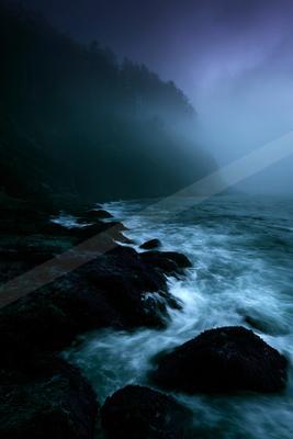 Dark rocky shoreline with waves crashing against boulders under a foggy, purple-hued sky, with a forested cliff in the background.