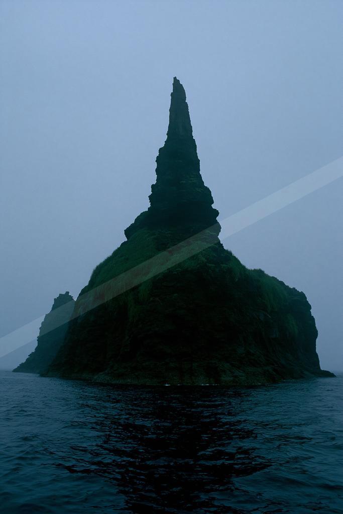 A tall, jagged sea stack covered in vibrant green moss rises from the dark ocean under a thick foggy sky.