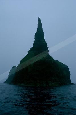 A tall, jagged sea stack covered in vibrant green moss rises from the dark ocean under a thick foggy sky.