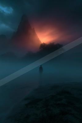 A lone silhouetted figure walks along a sandy path through thick blue fog toward a dramatic mountain peak illuminated by a glowing red and orange sunset or sunr