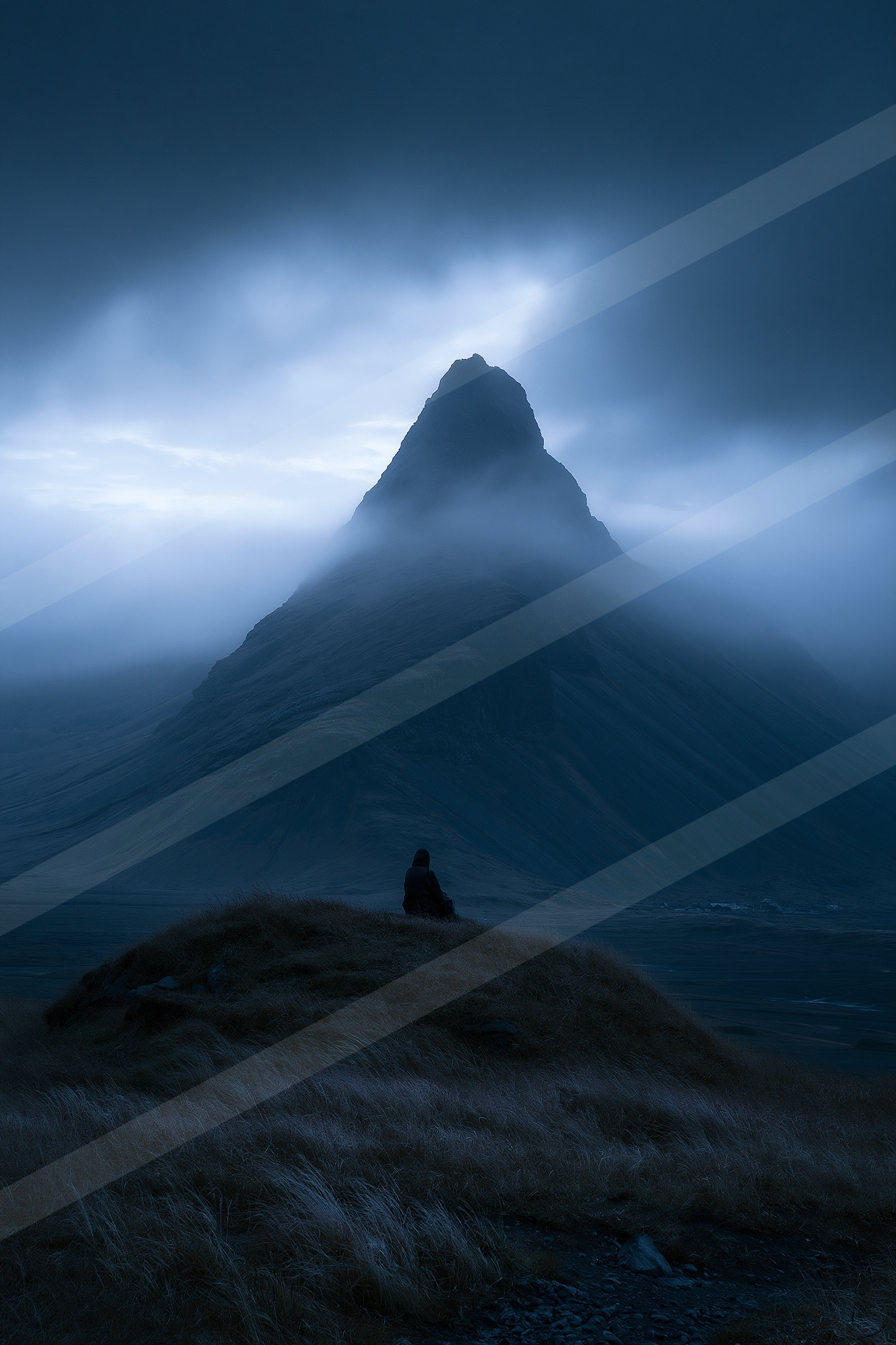 A lone figure sits on a grassy hill, silhouetted against the backdrop of a towering mountain peak shrouded in mist under a dark blue sky.