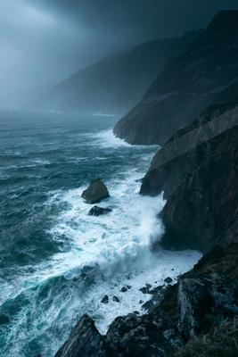 Powerful ocean waves with white foam crashing against dark rocky cliffs under a moody, overcast sky with mist and fog.