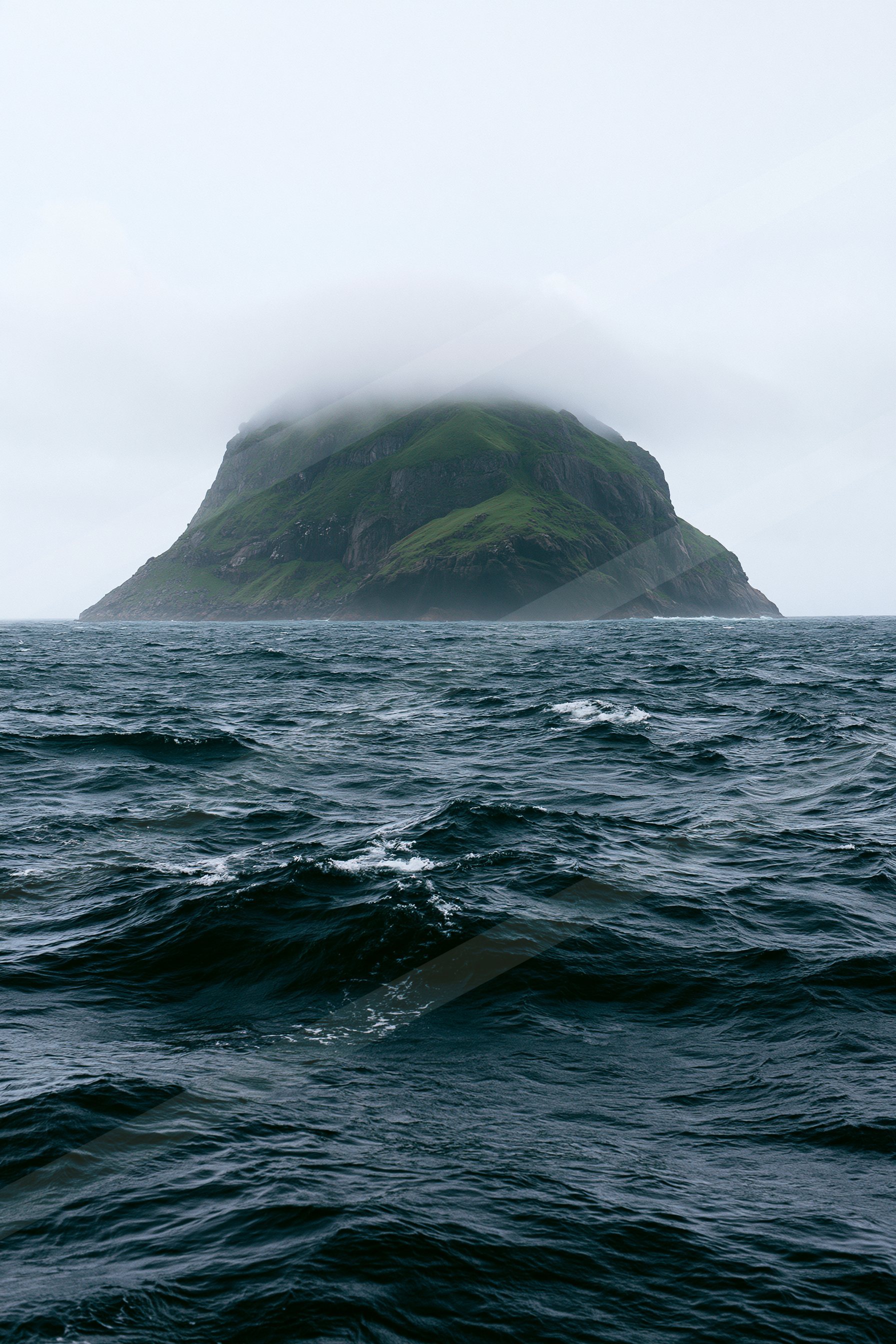 A solitary island covered in lush green vegetation rises from the ocean, partially shrouded by mist against a cloudy sky.