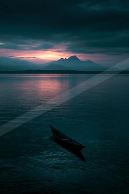 A wooden canoe floats alone on a vast calm lake reflecting the pink and purple hues of a dramatic sunset, with dark mountain silhouettes in the background under