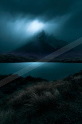 A dramatic, dark mountain peak shrouded in thick blue mist rises behind a calm lake under a stormy night sky, with tufts of grass in the foreground.