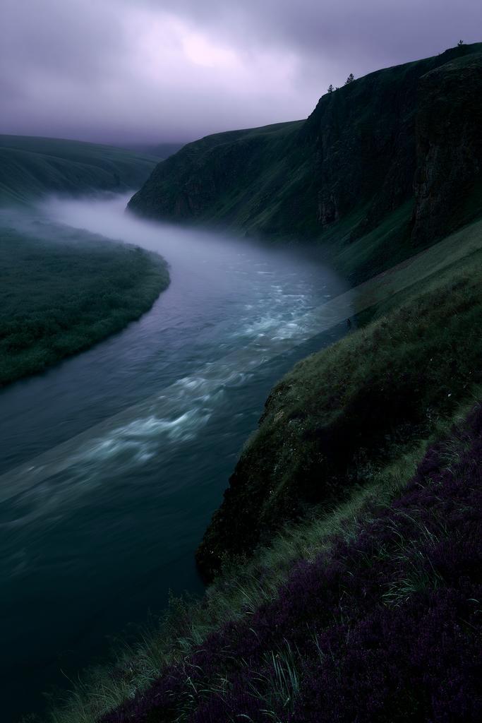 A winding river flows through a misty green valley between steep hills under a purple overcast sky, with purple wildflowers in the foreground.