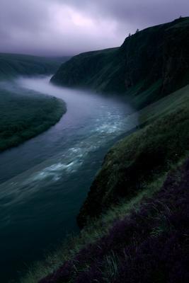 A winding river flows through a misty green valley between steep hills under a purple overcast sky, with purple wildflowers in the foreground.
