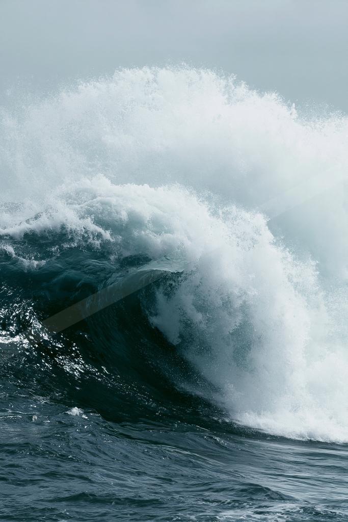 A massive ocean wave curling with a crest of white foam and spray against a muted sky.