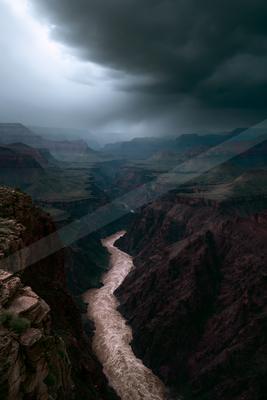 Moody aerial view of a winding muddy river carving through a vast, layered rock canyon under heavy, dark storm clouds with dramatic light breaking through.