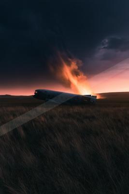 An abandoned airplane rests on a grassy field under a vivid sunset sky filled with dark clouds and fiery hues.