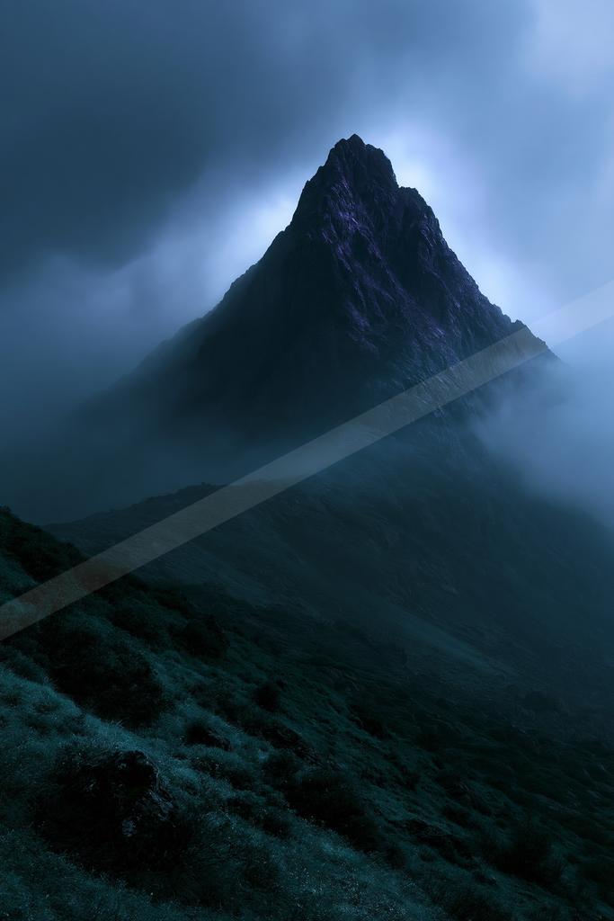 A sharp rocky mountain peak rises above thick rolling fog and low clouds under a dark blue twilight sky, with a grassy hillside in the foreground.
