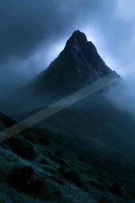 A sharp rocky mountain peak rises above thick rolling fog and low clouds under a dark blue twilight sky, with a grassy hillside in the foreground.