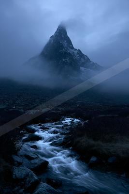 A sharp rocky mountain peak rises through thick fog above a fast-flowing stream cascading over rocks in a dark, moody alpine valley.
