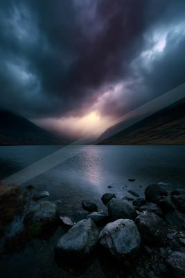 Moody landscape with dark storm clouds over a calm mountain lake, rocky shoreline in foreground and light breaking through the clouds in the distance.