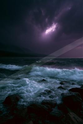 Powerful ocean waves crash against dark rocks under a dramatic purple stormy sky illuminated by a bright lightning bolt.