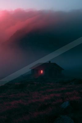 A solitary wooden cabin with a single glowing red window stands on a grassy hillside, surrounded by thick, colorful pink and blue fog rolling over dark mountain