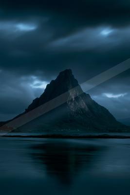 A steep rocky mountain peak rises against a dark, cloudy night sky, its reflection visible in the calm foreground water.