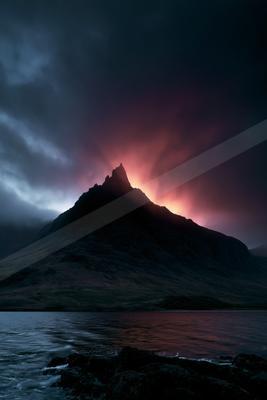 A sharp rocky mountain peak rises above a body of water under a dramatic twilight sky filled with deep red, purple, and dark blue clouds.