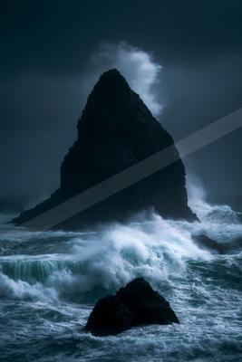 A tall dark sea stack rises from turbulent ocean waters as powerful white waves crash against it under a stormy sky.