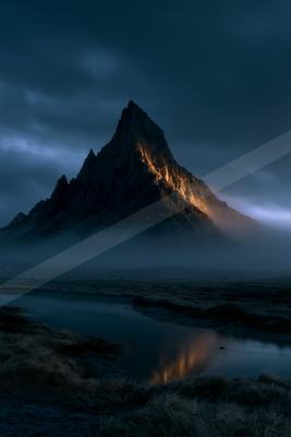 A sharp rocky mountain peak catches warm light on its ridge against a dark cloudy twilight sky, with low mist surrounding its base and a calm lake in the foregr