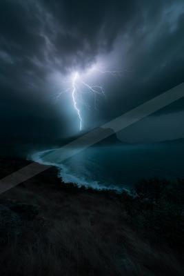 A powerful lightning bolt illuminates the dark sky over a coastal landscape, highlighting the ocean waves and silhouetted hills beneath stormy clouds.