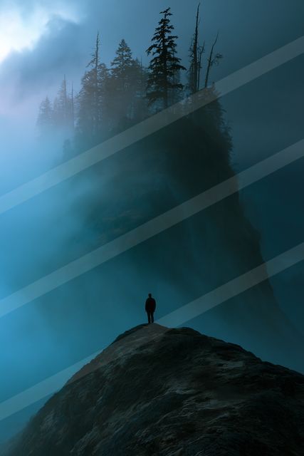 A lone figure stands silhouetted atop a rugged mountain peak shrouded in atmospheric fog and surrounded by tall evergreen trees.